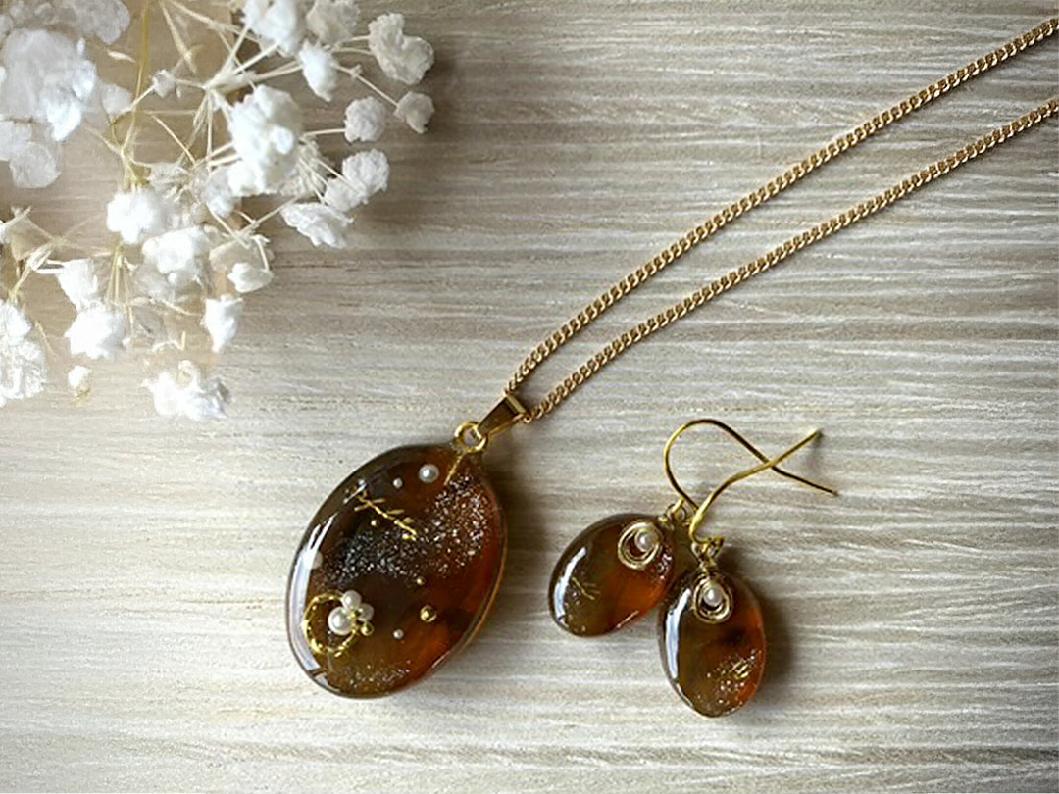 琥珀のような深みのあるブラウンカラーが美しいレジンで仕立てたネックレスとピアスのセット作品。透明感の中に繊細なラメや小粒パール、ゴールドパーツが浮かび、光を受けるたび上品にきらめきます。落ち着いた色合いながら華やかさもあり幅広く寄り添ってくれるアクセ。