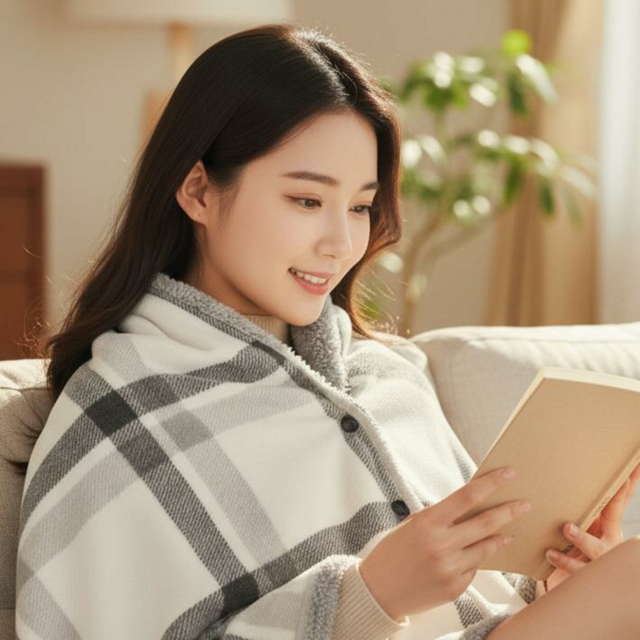 グレーチェック柄のフリース3WAYブランケットを羽織り、ソファで読書を楽しむ女性。やわらかく軽い素材で肩から膝まで暖かく包み、おうち時間や冷え対策にぴったりの冬の人気アイテムです。